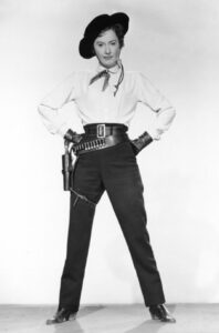 black and white photograph of Barbara Stanwyck in cowboy outfit: hat, holster, pants, gunbelt, boots.