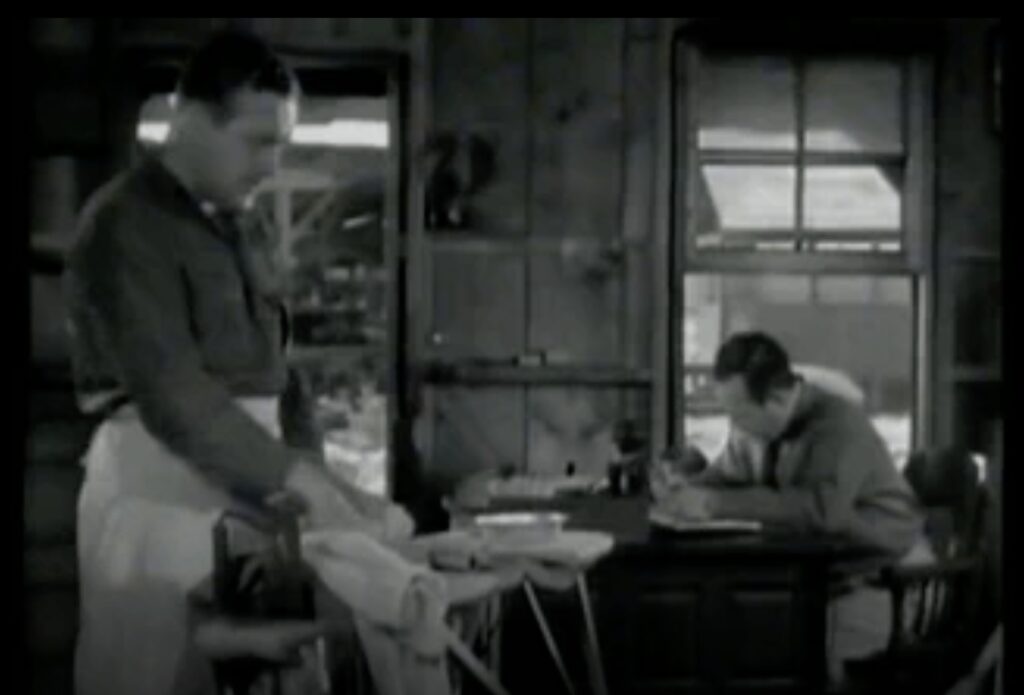 the Gay cook also does the laundry, ironing clothing, while an officer sits at a table with papers.
