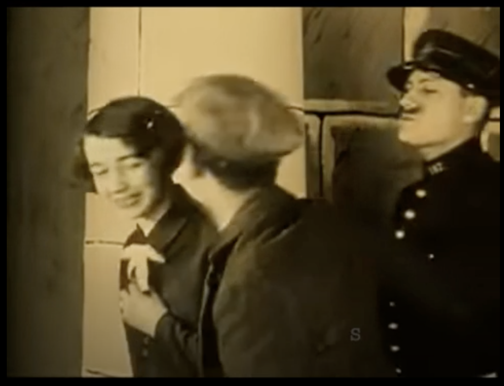 still from "Invitation to a Journey" - a woman with a false mustache acts as a cop, standing behind a young couple