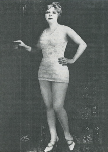 photograph of Mae West in short dress, heels, hand on hip, other hand resting on a shelf, short hair, made up face.