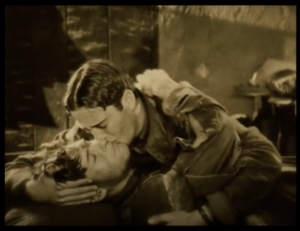 still from the film "Wings" - two men, soldiers, embrace and kiss, one of them lying injured, the other comforting him.