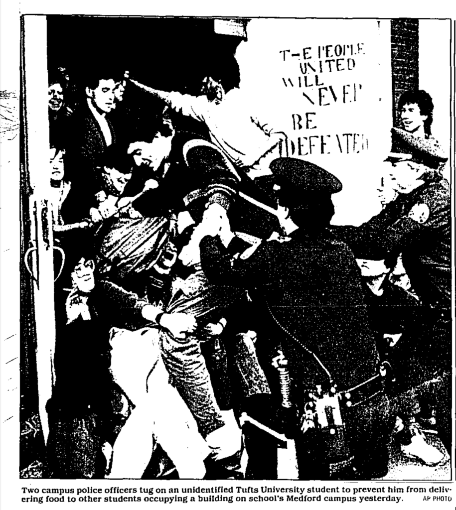 newspaper photograph of police trying to pull students away from other students, in a doorway, a sign reads "The People united will never be defeated" - caption: "Two campus police officers tug on an unidentified Tufts University student to prevent him from delivering food to other students occupying a building on school's Medford campus"