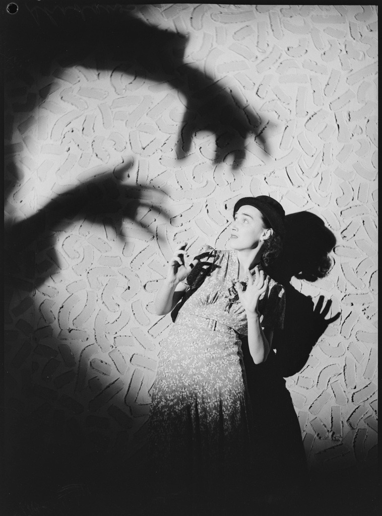 old black and white staged picture: a woman stands against a wall, a huge shadow over her of hands reaching towards her ominously